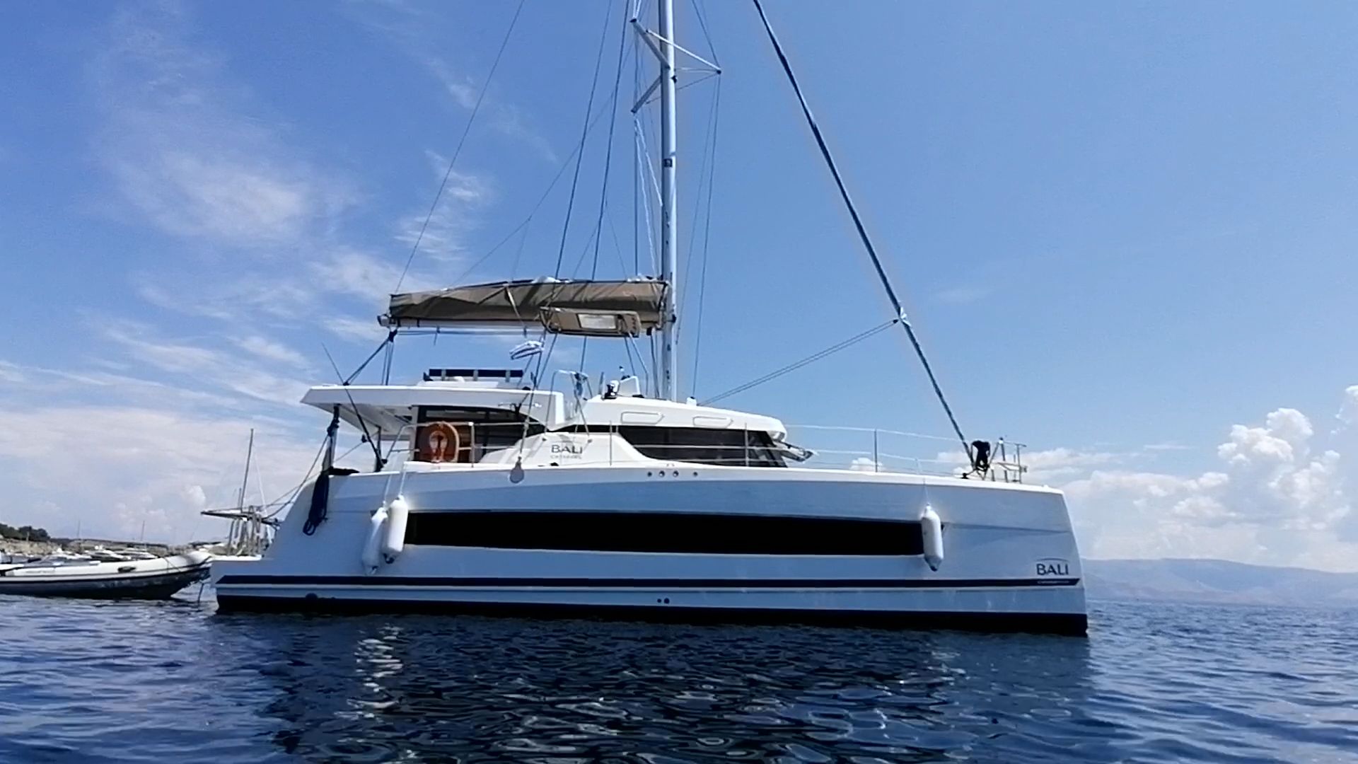 A white catamaran is shown floating on blue water under a clear sky with scattered clouds. Mountains are visible in the distance.